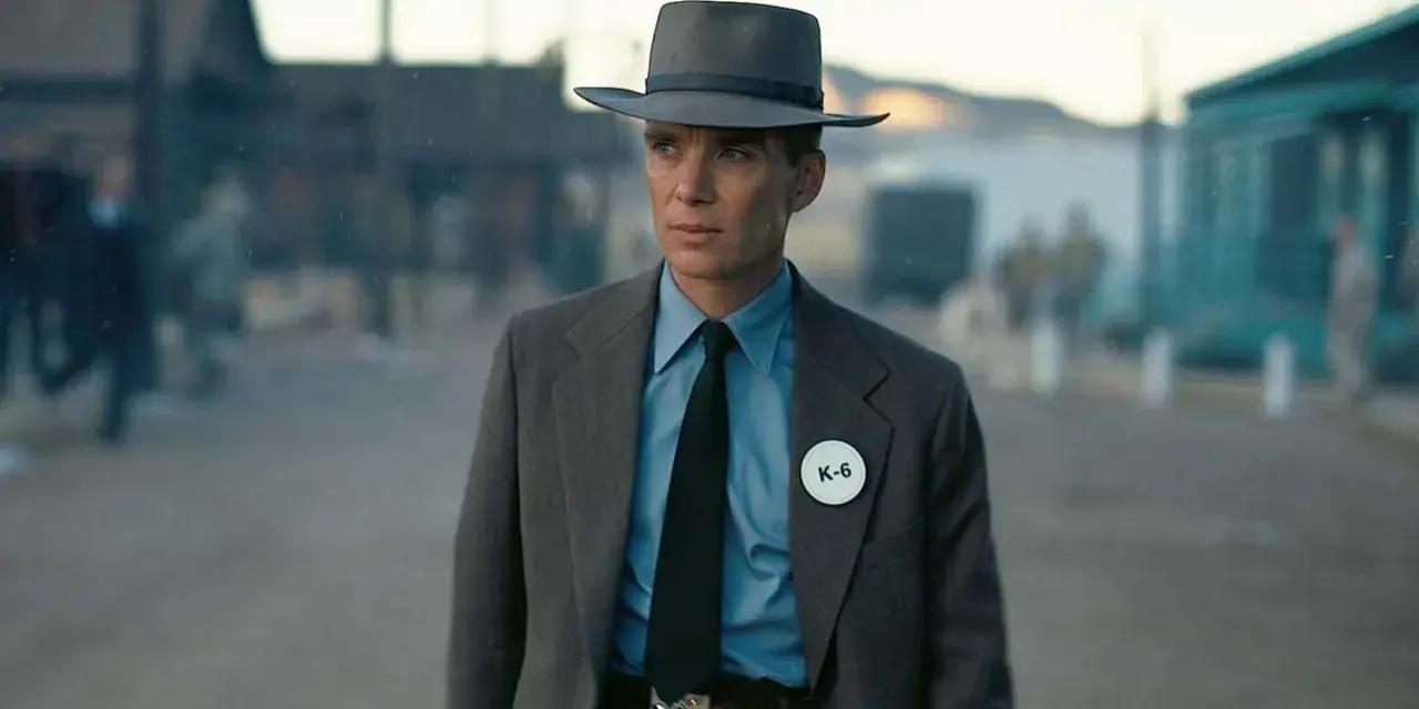Julius Robert Oppenheimer w kapeluszu i garniturze, z odznaką K-6, w scenerii przypominającej miasteczko.