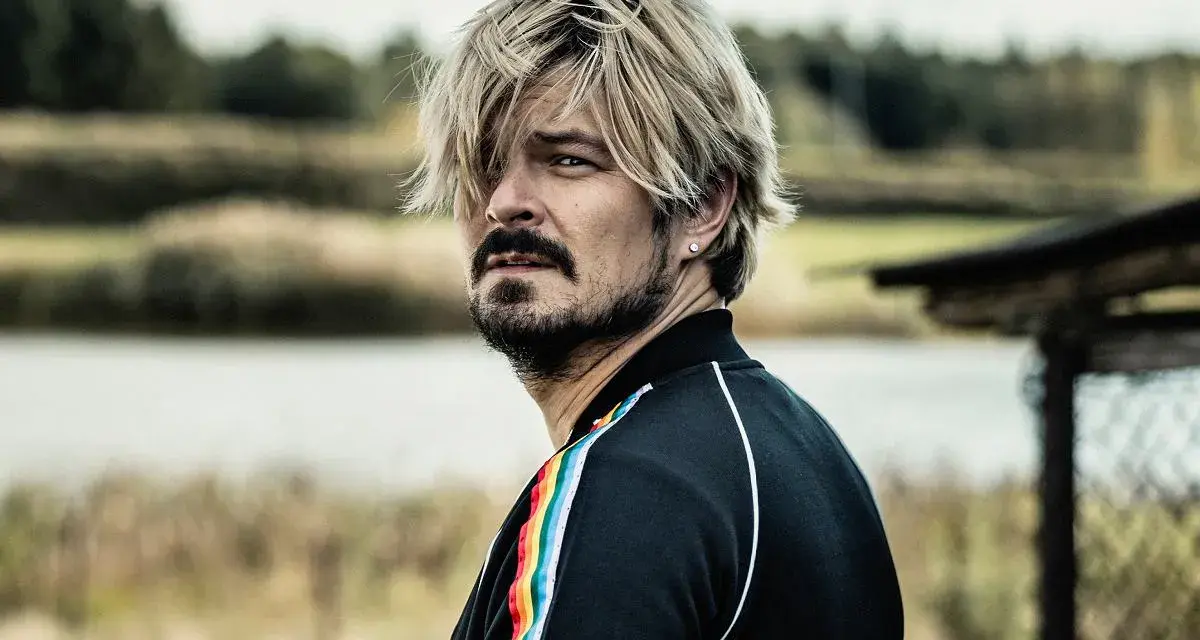 Mężczyzna z blond włosami i zarostem, w czarnej bluzie z tęczowym lampasem, spogląda w bok, jakby mierzył się ze wszystkimi naszymi strachami.