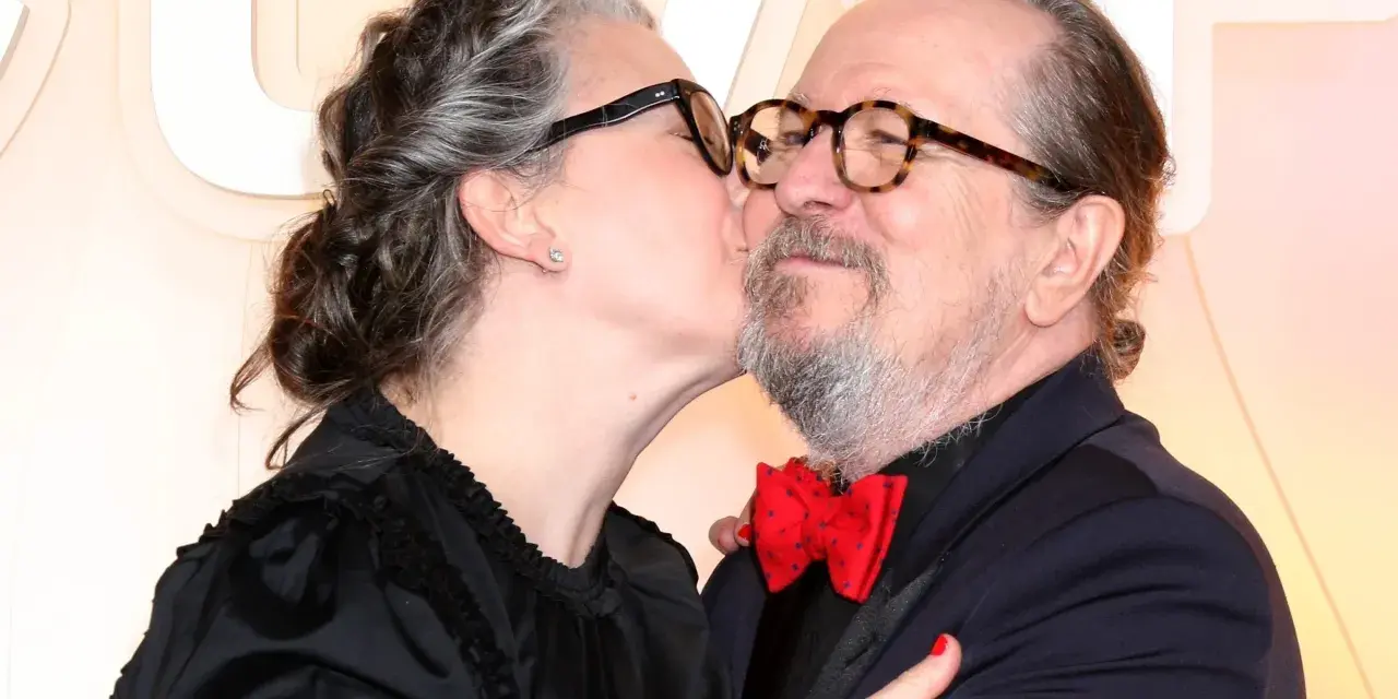 Gary Oldman otrzymuje czuły pocałunek od swojej żony. Oboje mają okulary, a ona nosi czarną sukienkę.