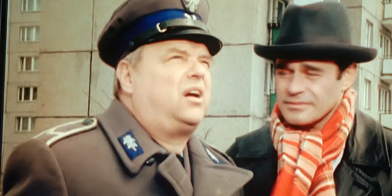 Dwóch mężczyzn w kapeluszach, jeden w mundurze, drugi w szaliku. Brunet wieczorową porą, w czarnym kapeluszu i czerwonym szaliku, rozmawia z policjantem.