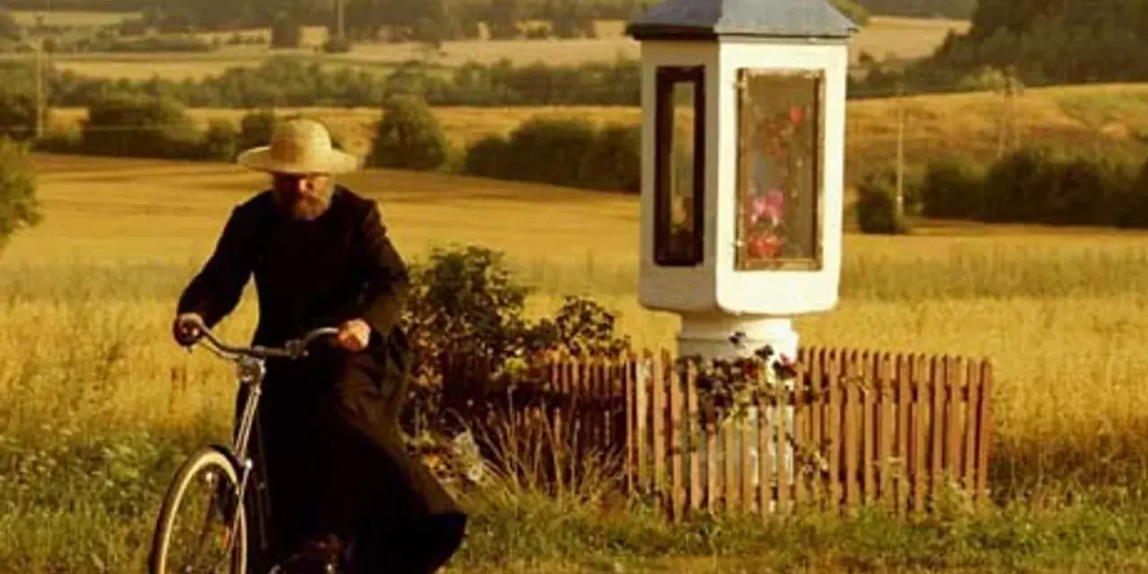 Mężczyzna w słomkowym kapeluszu i czarnej sutannie jedzie rowerem przez pola, mijając kapliczkę. Wydaje się, że jest u pana boga za piecem.