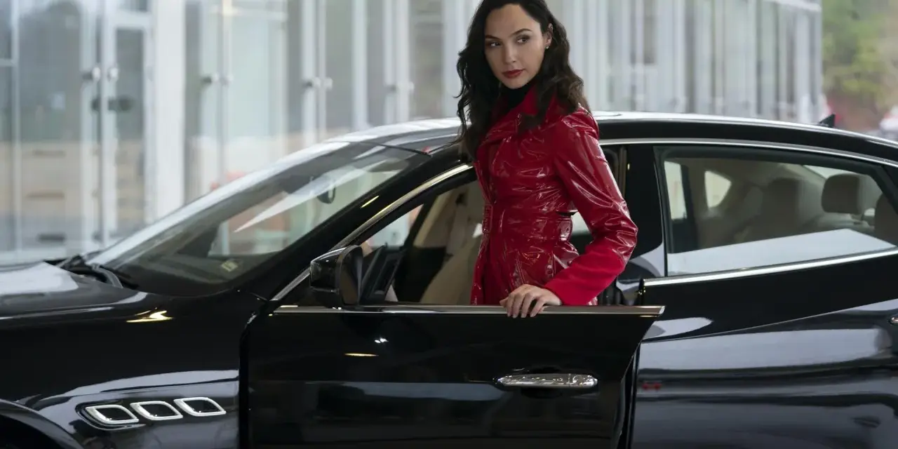 Kobieta w błyszczącym, czerwonym płaszczu opiera się o czarne auto. Wygląda jak z filmu, może to jej czerwona nota 2?