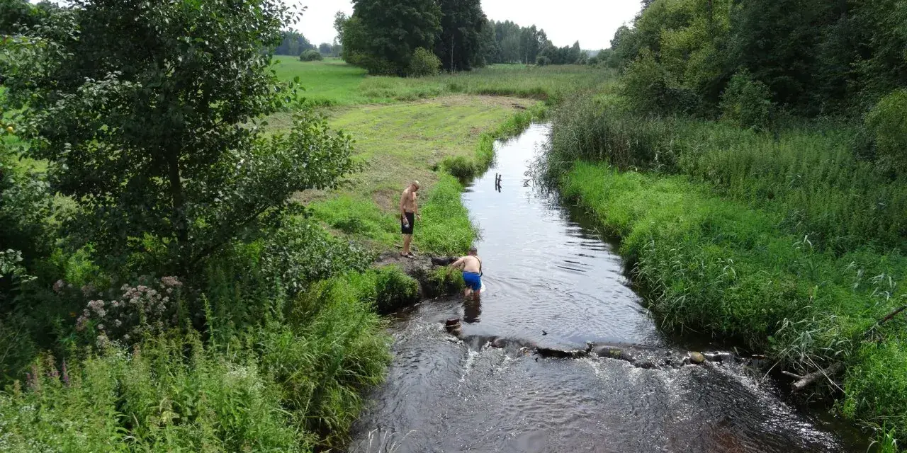 Dwóch mężczyzn w kąpielówkach pluska się w rzece, czując się jak u pana boga w ogródku. Otacza ich bujna zieleń i spokój natury.