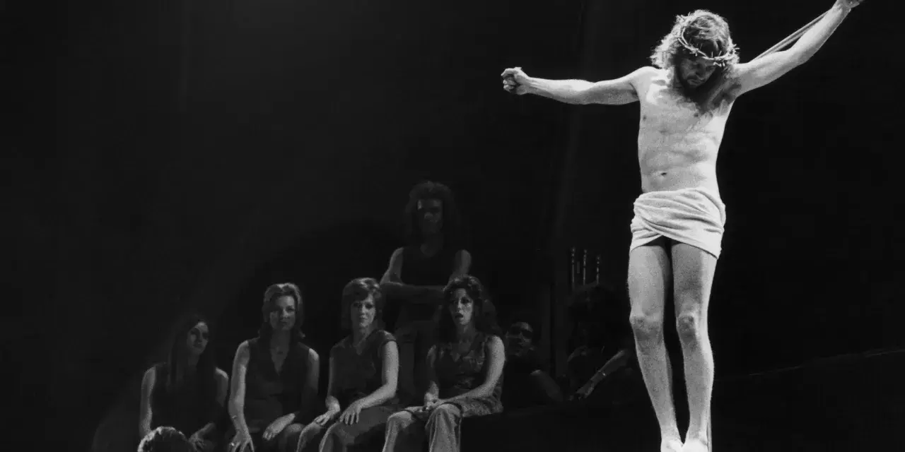 Scena z musicalu Jesus Christ Superstar: Jezus na krzyżu, otoczony przez postaci, w tym kobiety siedzące wpatrzone w niego.