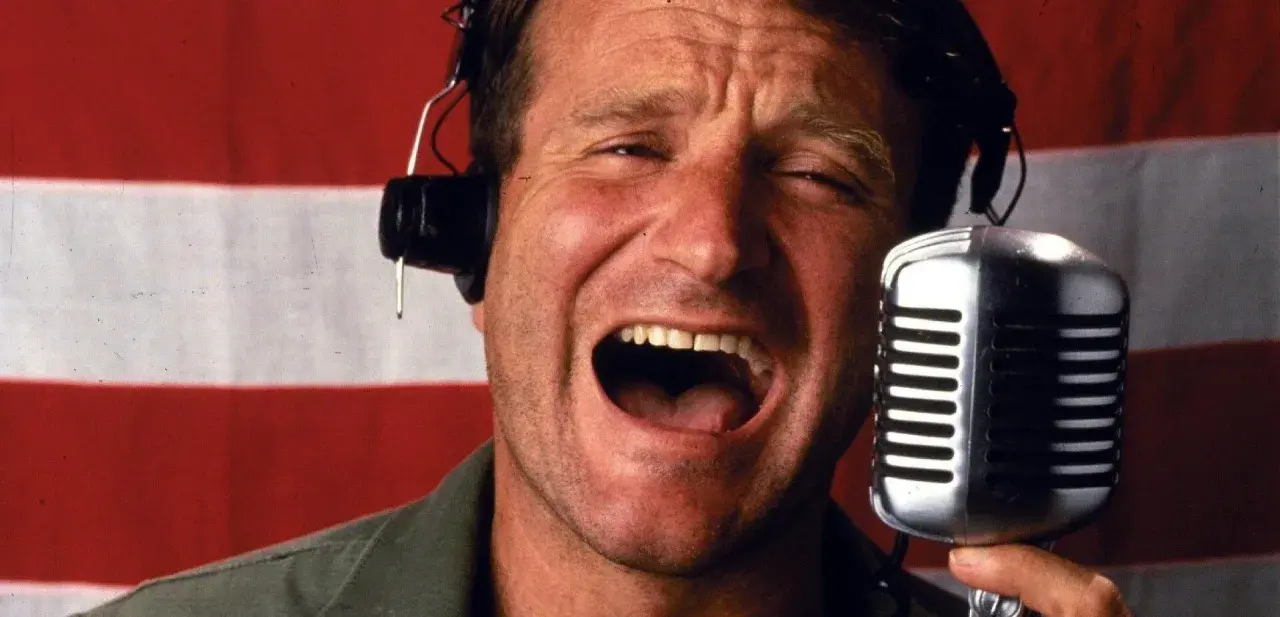 Robin Williams z mikrofonem i słuchawkami na tle flagi. W jakich filmach grał Robin Williams?