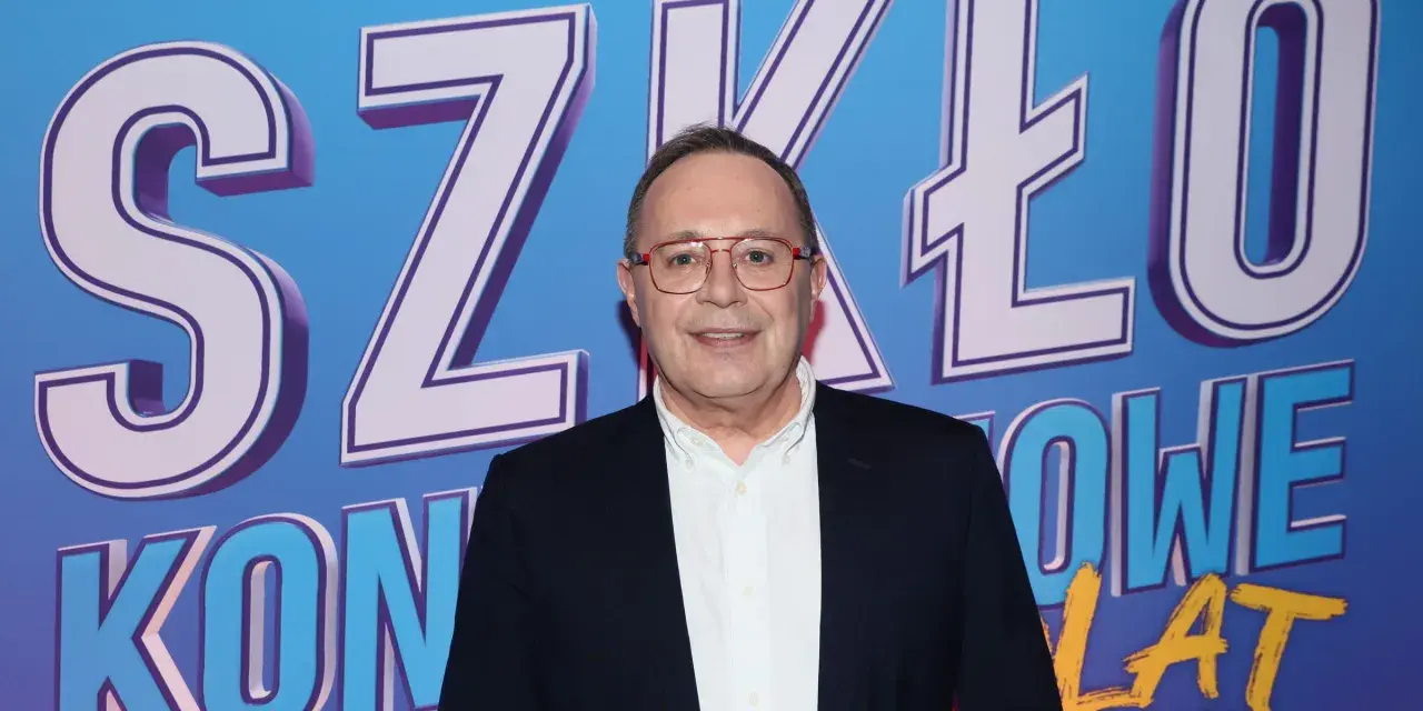 Uśmiechnięty mężczyzna w okularach na tle napisu "SZKŁO KONTAKTOWE". Prowadzący wydarzenie, gotowy do rozmowy.