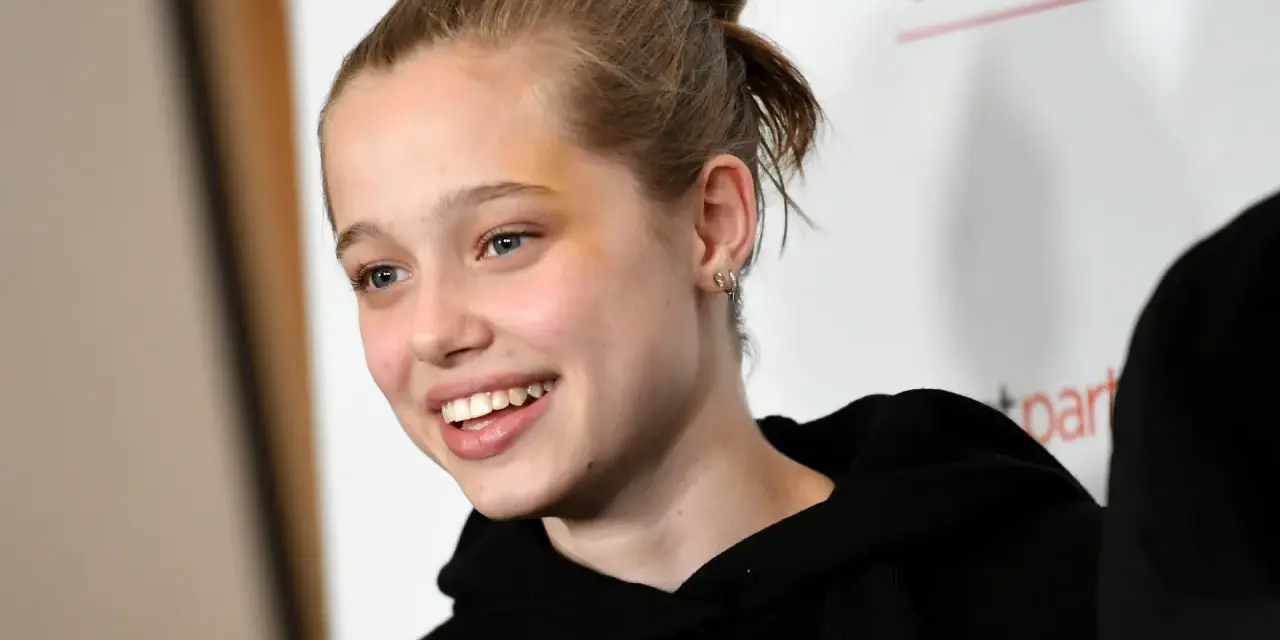 Uśmiechnięta Shiloh Jolie Pitt z włosami upiętymi w kok, w czarnej bluzie z kapturem.