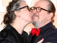Gary Oldman otrzymuje czuły pocałunek od swojej żony. Oboje mają okulary, a ona nosi czarną sukienkę.