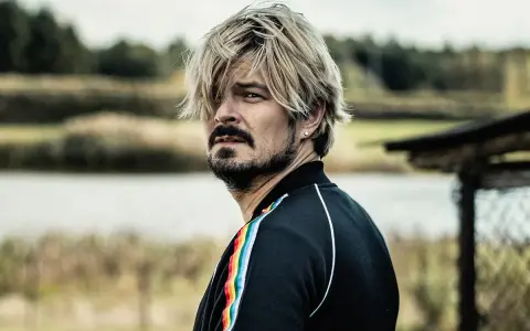 Mężczyzna z blond włosami i zarostem, w czarnej bluzie z tęczowym lampasem, spogląda w bok, jakby mierzył się ze wszystkimi naszymi strachami.