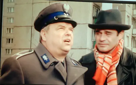 Dwóch mężczyzn w kapeluszach, jeden w mundurze, drugi w szaliku. Brunet wieczorową porą, w czarnym kapeluszu i czerwonym szaliku, rozmawia z policjantem.