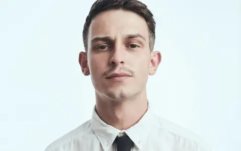 Młody mężczyzna, Vito Bambino, w białej koszuli i czarnym krawacie. Jego wzrost jest imponujący, a spojrzenie intensywne.