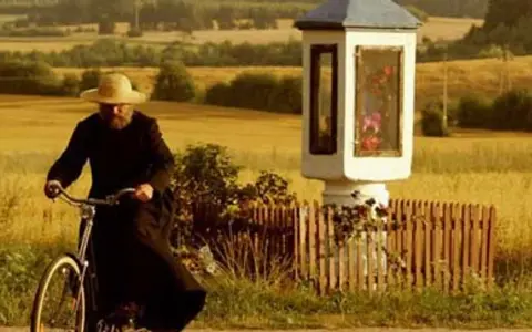 Mężczyzna w słomkowym kapeluszu i czarnej sutannie jedzie rowerem przez pola, mijając kapliczkę. Wydaje się, że jest u pana boga za piecem.