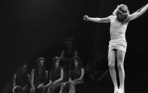 Scena z musicalu Jesus Christ Superstar: Jezus na krzyżu, otoczony przez postaci, w tym kobiety siedzące wpatrzone w niego.