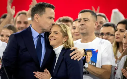 Rafał Trzaskowski, choć nie znamy jego dokładnego wieku, czule całuje żonę na tle tłumu.