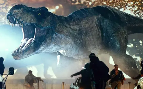 Gigantyczny Tyranozaur Rex z Jurassic World 3 z otwartą paszczą ściga ludzi wśród samochodów.