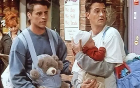 Chandler i Joey z dzieckiem i misiem w nosidełku. Chandler wygląda na lekko zaniepokojonego.