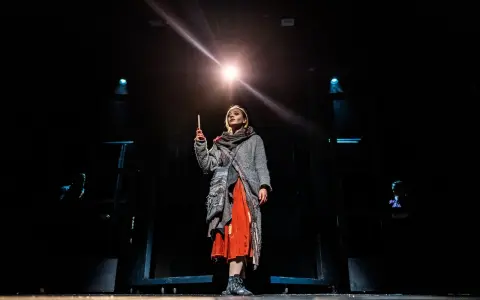 Dziewczynka z zapałkami na scenie, trzymająca płonącą zapałkę, w otoczeniu mroku i teatralnego światła.