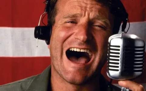 Robin Williams z mikrofonem i słuchawkami na tle flagi. W jakich filmach grał Robin Williams?