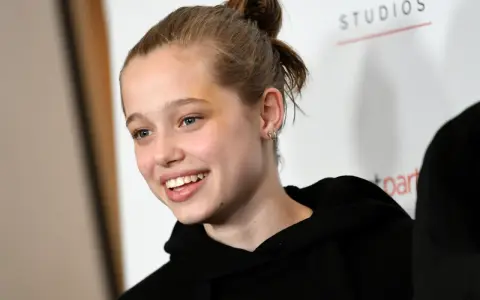 Uśmiechnięta Shiloh Jolie Pitt z włosami upiętymi w kok, w czarnej bluzie z kapturem.