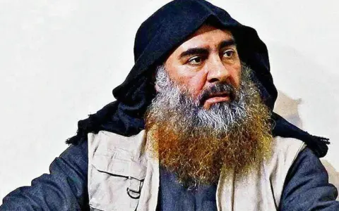 Mężczyzna z długą, siwiejącą brodą i czarnym nakryciem głowy, przypominający Abu Bakra al-Baghdadiego, siedzi w jasnym ubraniu.