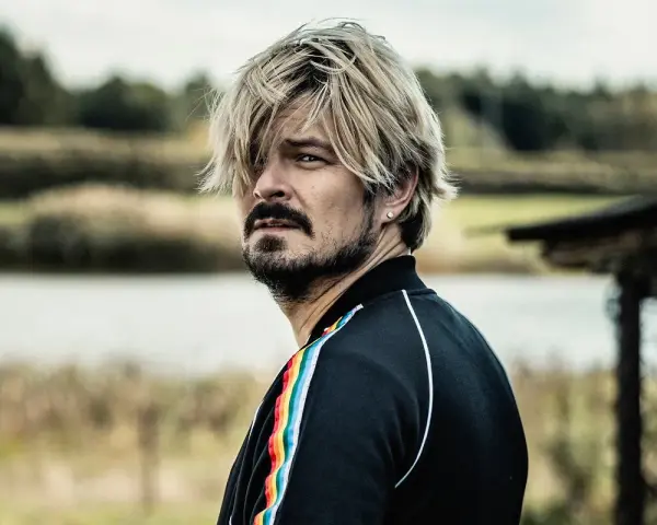 Mężczyzna z blond włosami i zarostem, w czarnej bluzie z tęczowym lampasem, spogląda w bok, jakby mierzył się ze wszystkimi naszymi strachami.