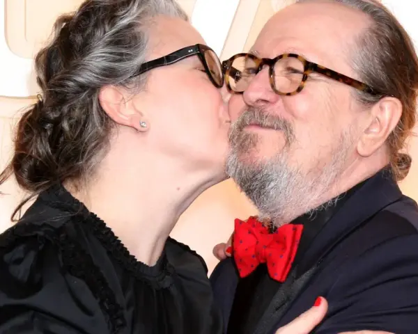 Gary Oldman otrzymuje czuły pocałunek od swojej żony. Oboje mają okulary, a ona nosi czarną sukienkę.