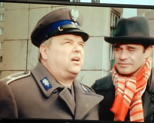 Dwóch mężczyzn w kapeluszach, jeden w mundurze, drugi w szaliku. Brunet wieczorową porą, w czarnym kapeluszu i czerwonym szaliku, rozmawia z policjantem.