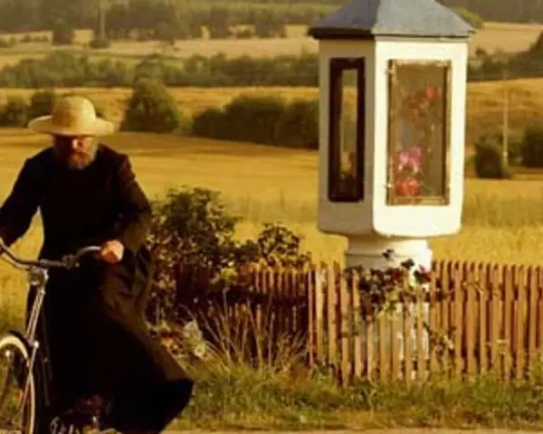 Mężczyzna w słomkowym kapeluszu i czarnej sutannie jedzie rowerem przez pola, mijając kapliczkę. Wydaje się, że jest u pana boga za piecem.