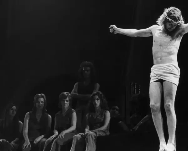 Scena z musicalu Jesus Christ Superstar: Jezus na krzyżu, otoczony przez postaci, w tym kobiety siedzące wpatrzone w niego.