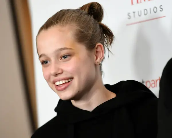 Uśmiechnięta Shiloh Jolie Pitt z włosami upiętymi w kok, w czarnej bluzie z kapturem.