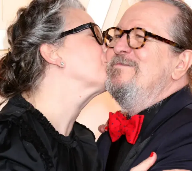 Gary Oldman otrzymuje czuły pocałunek od swojej żony. Oboje mają okulary, a ona nosi czarną sukienkę.