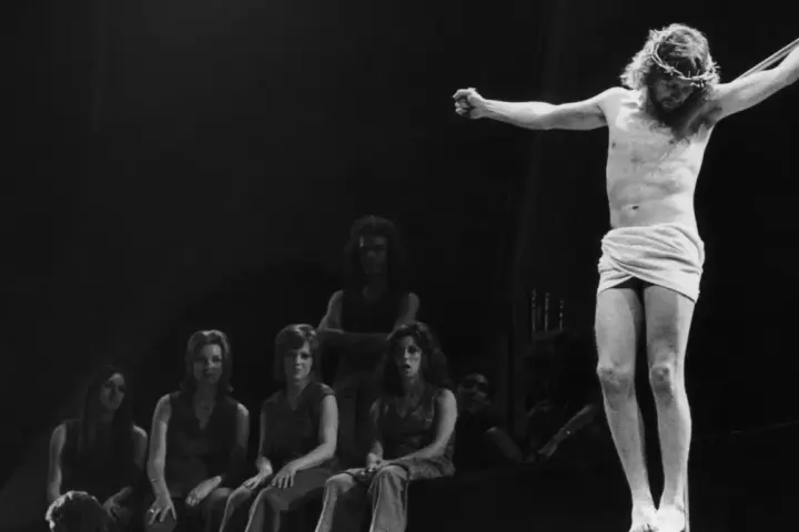 Scena z musicalu Jesus Christ Superstar: Jezus na krzyżu, otoczony przez postaci, w tym kobiety siedzące wpatrzone w niego.