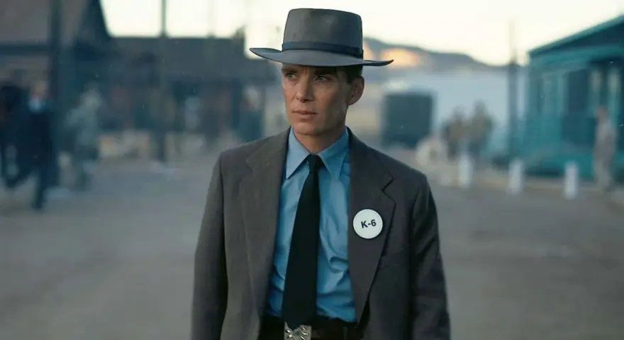 Julius Robert Oppenheimer w kapeluszu i garniturze, z odznaką K-6, w scenerii przypominającej miasteczko.