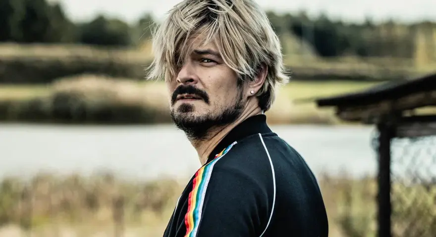 Mężczyzna z blond włosami i zarostem, w czarnej bluzie z tęczowym lampasem, spogląda w bok, jakby mierzył się ze wszystkimi naszymi strachami.