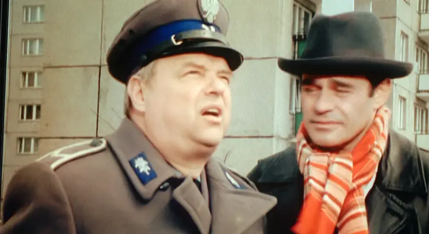 Dwóch mężczyzn w kapeluszach, jeden w mundurze, drugi w szaliku. Brunet wieczorową porą, w czarnym kapeluszu i czerwonym szaliku, rozmawia z policjantem.