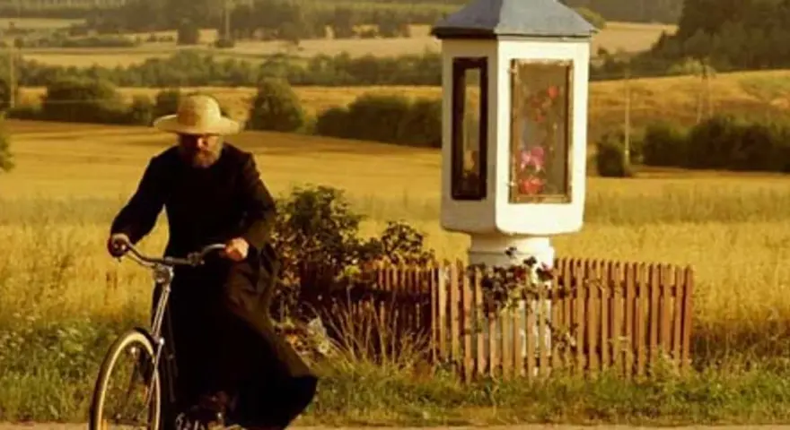 Mężczyzna w słomkowym kapeluszu i czarnej sutannie jedzie rowerem przez pola, mijając kapliczkę. Wydaje się, że jest u pana boga za piecem.