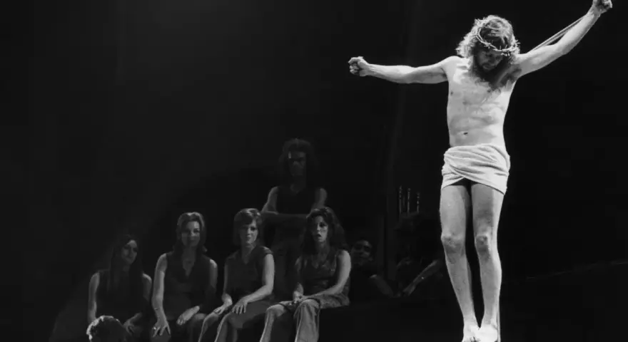 Scena z musicalu Jesus Christ Superstar: Jezus na krzyżu, otoczony przez postaci, w tym kobiety siedzące wpatrzone w niego.