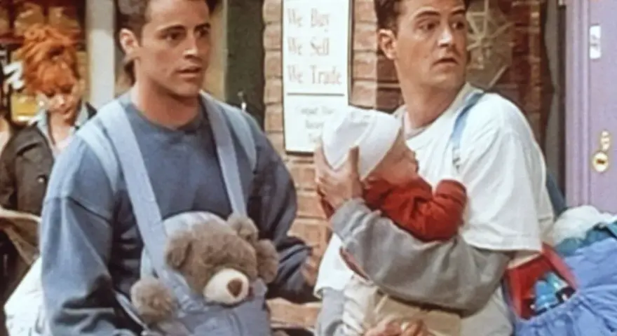Chandler i Joey z dzieckiem i misiem w nosidełku. Chandler wygląda na lekko zaniepokojonego.