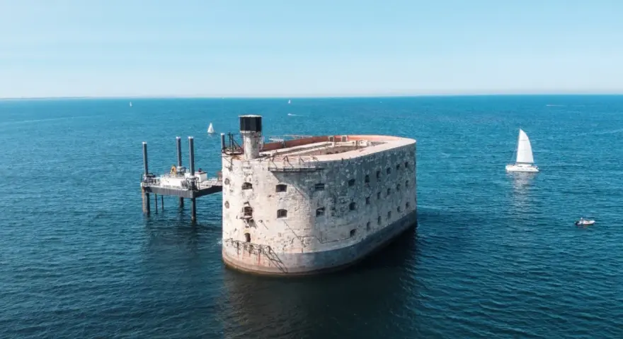 Fort Boyard, potężna twierdza na morzu, otoczona błękitną wodą. Obok platforma wiertnicza i żaglówka.