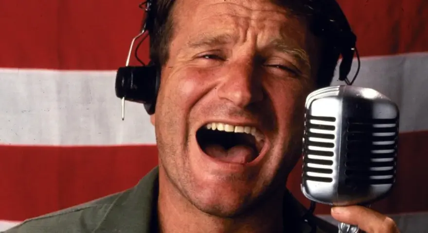 Robin Williams z mikrofonem i słuchawkami na tle flagi. W jakich filmach grał Robin Williams?