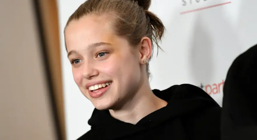 Uśmiechnięta Shiloh Jolie Pitt z włosami upiętymi w kok, w czarnej bluzie z kapturem.