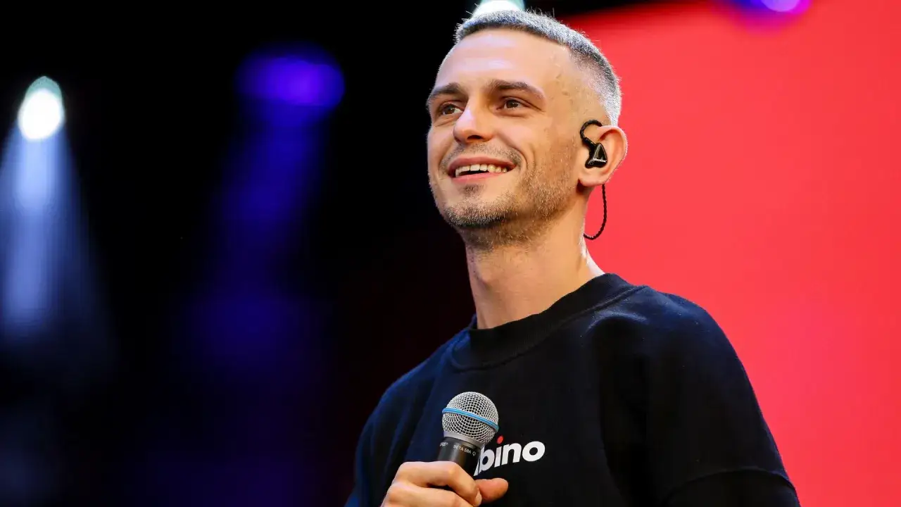 Uśmiechnięty Vito Bambino na scenie, z mikrofonem w dłoni. Jego wzrost jest imponujący, a na szyi widać napis 