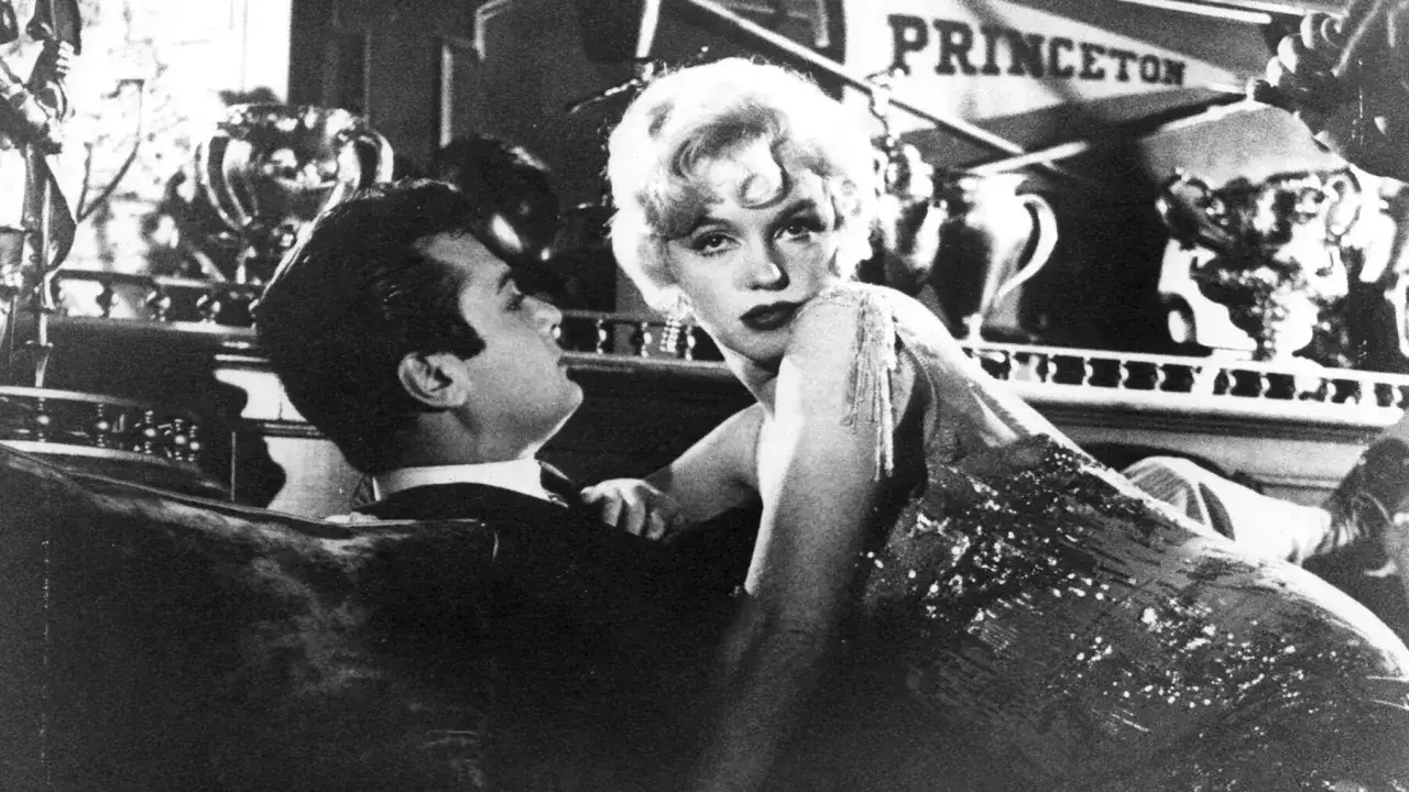 Marilyn Monroe uwodzi Tony'ego Curtisa, pół żartem pół serio, na tle napisu 