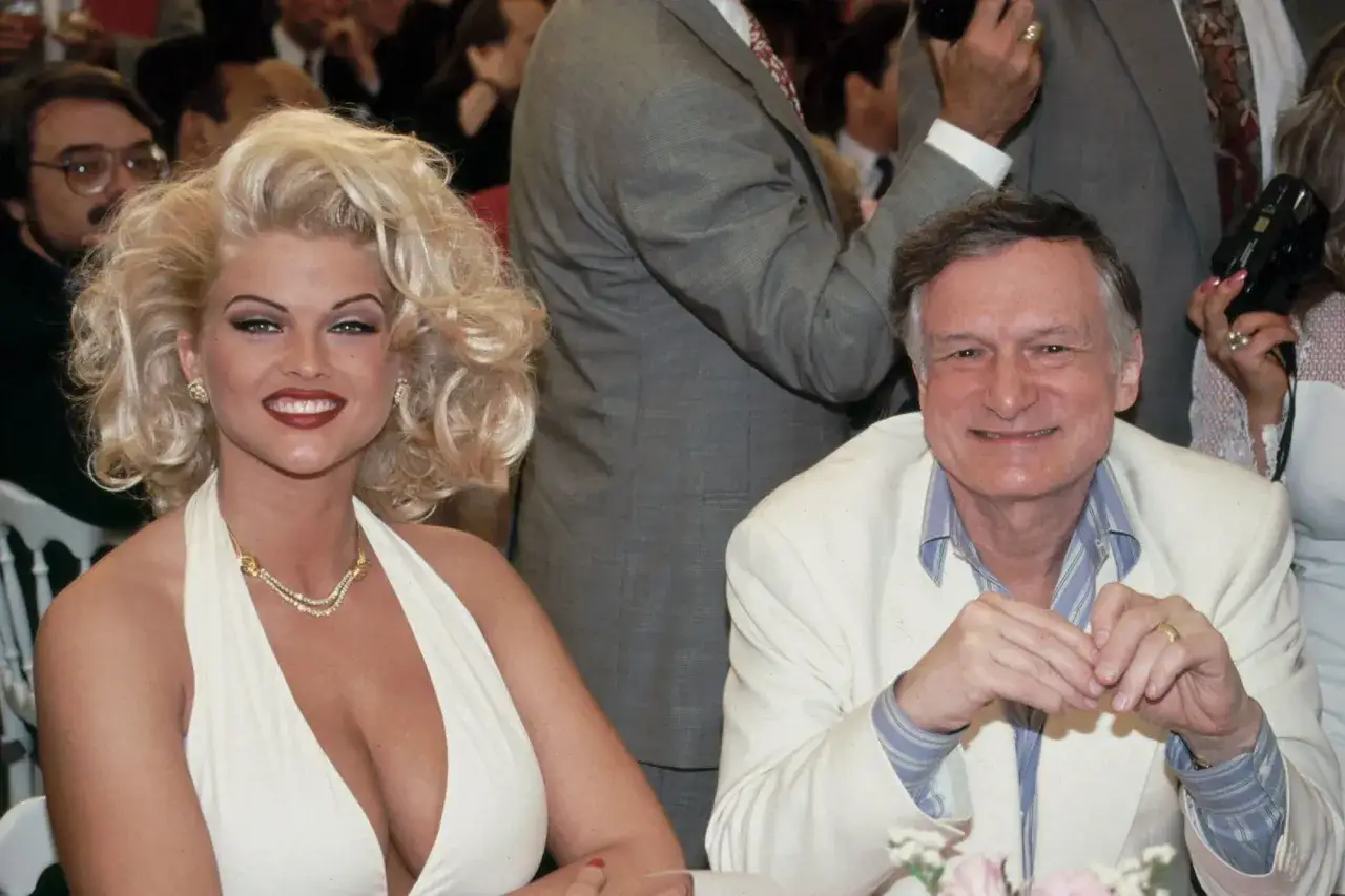 Anna Nicole Smith w białej sukni i Hugh Hefner w jasnej marynarce uśmiechają się do zdjęcia.