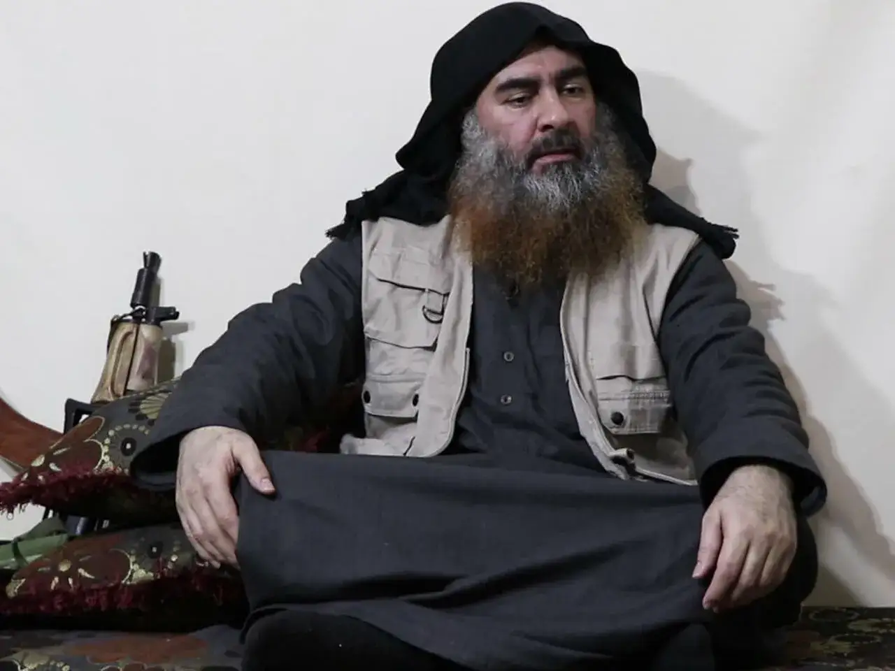 Abu Bakr al-Baghdadi, z brodą i w czarnym nakryciu głowy, siedzi w szarym ubraniu i kamizelce. Obok niego leży karabin.