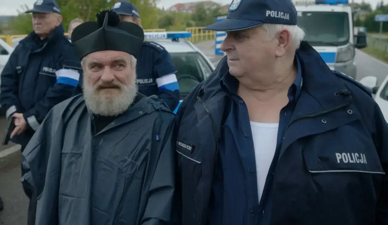Ksiądz z brodą i policjant rozmawiają na tle radiowozów. Wygląda, jakby byli u pana Boga w ogródku, w środku akcji.