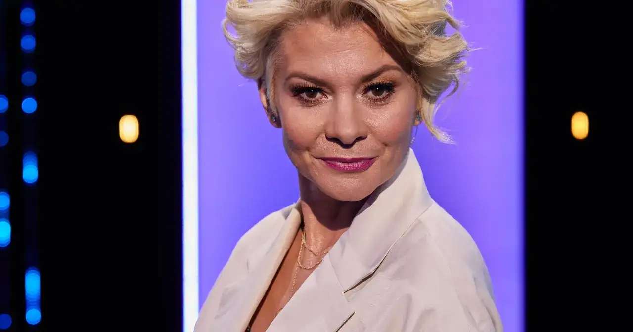 Beata Olga Kowalska z uśmiechem na twarzy, w jasnej marynarce, na tle fioletowo-czarnej sceny z niebieskimi i żółtymi światłami.
