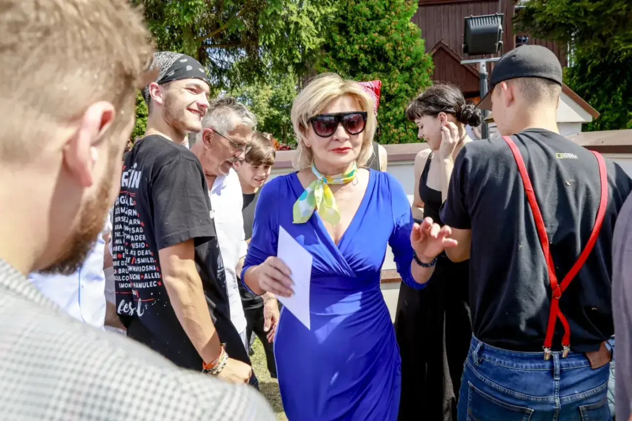 Beata Olga Kowalska w niebieskiej sukience i kolorowym szalu, otoczona ludźmi, trzyma kartkę.