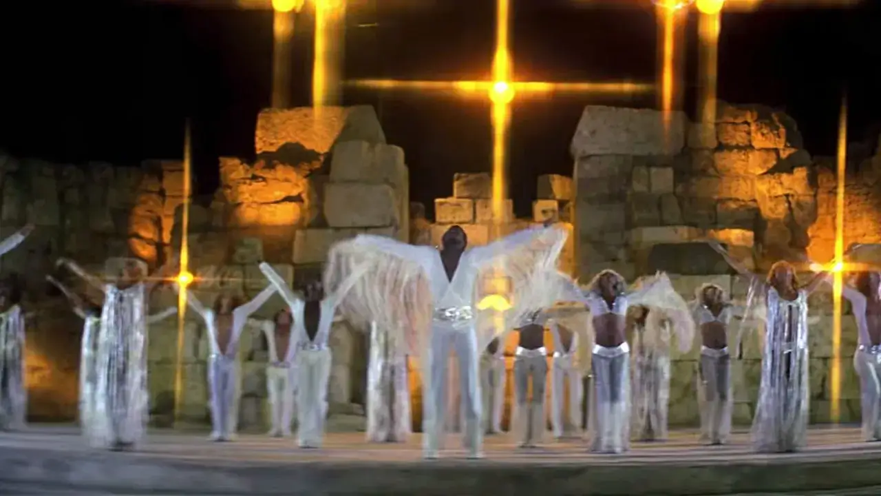 Scena z musicalu Jesus Christ Superstar. Tancerze w białych, błyszczących strojach unoszą ręce ku niebu, gdzie jaśnieją złote krzyże.