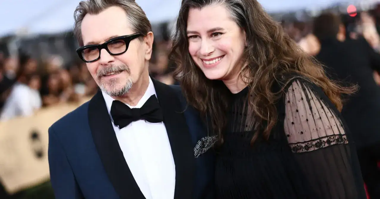 Gary Oldman z żoną, oboje uśmiechnięci, na czerwonym dywanie. On w eleganckim garniturze, ona w czarnej sukni.